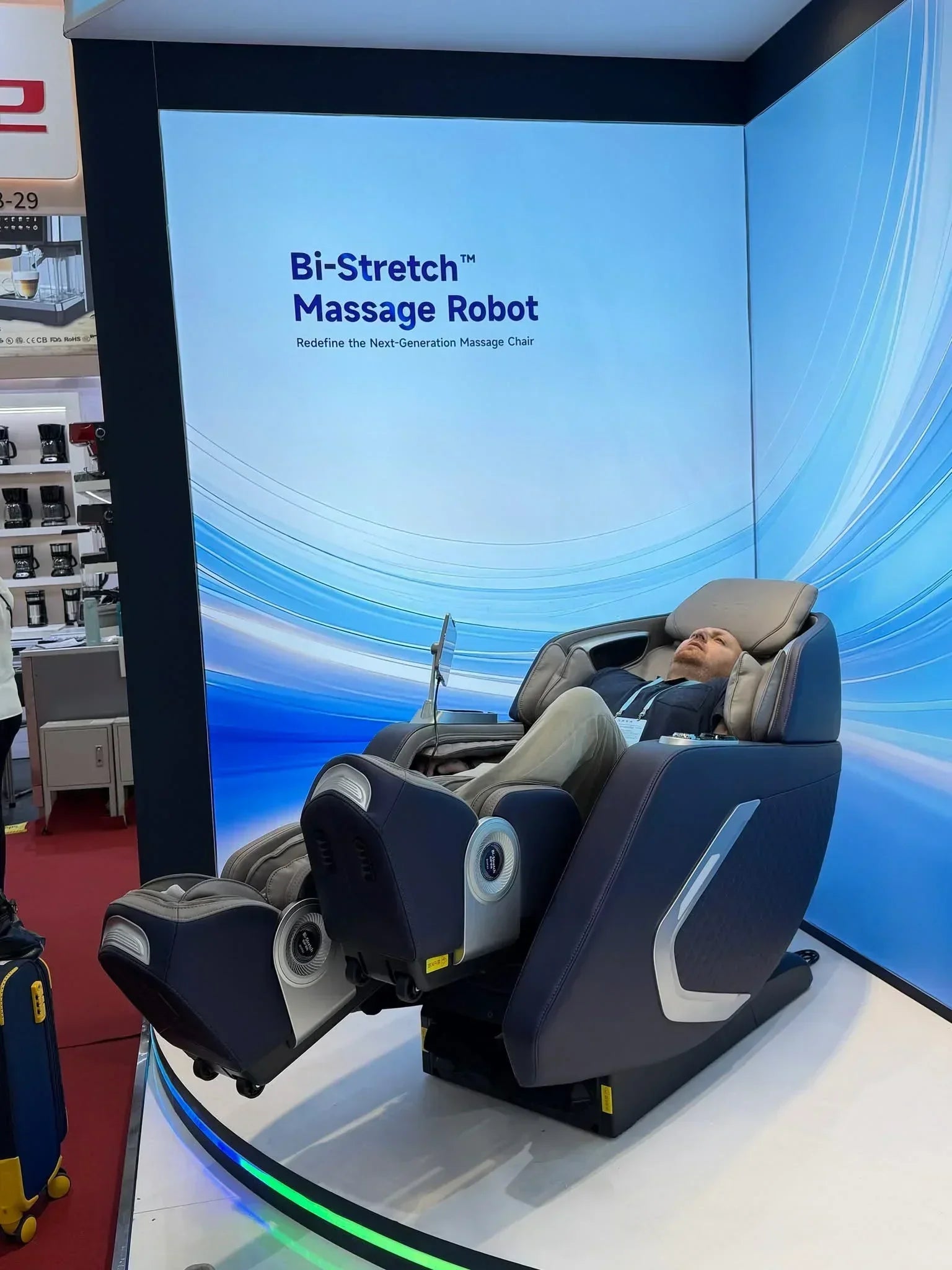 Man relaxing in a Bi-Stretch massage chair at an exhibition booth with modern blue backdrop